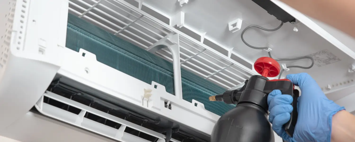 Hand wearing blue glove spraying cleaning solution inside wall-mounted air conditioner
